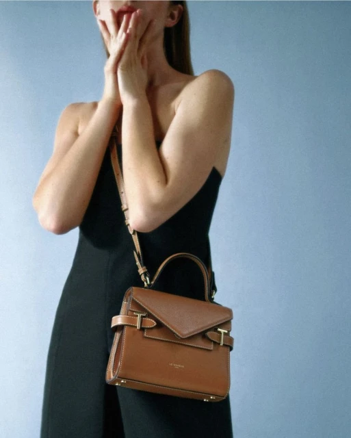 Adult woman with a surprised expression wearing a black jumpsuit poses with a brown leather satchel handbag against a blue background.