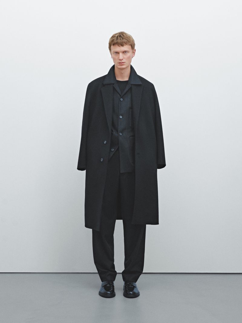 Full-length shot of a slender adult male fashion model in a black oversized coat, shirt, and pants on a grey studio floor against a white wall.