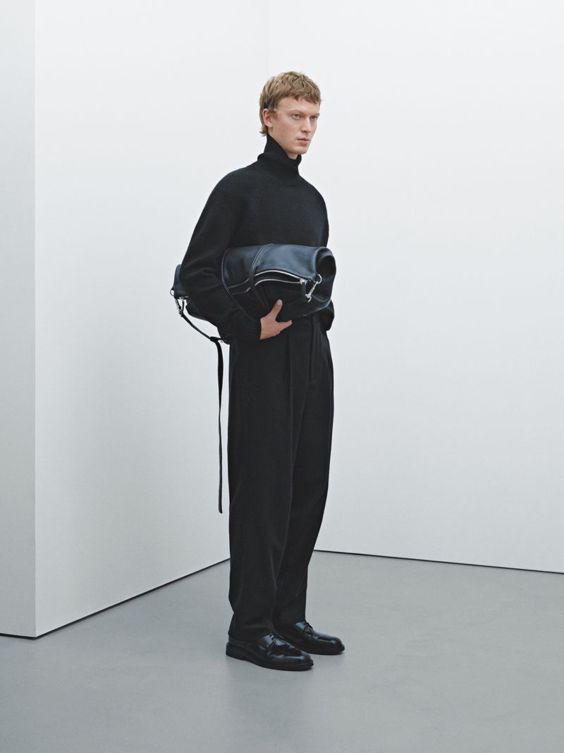 Male model in a black turtleneck and black pants holding a black leather bag in a white photo studio setting.