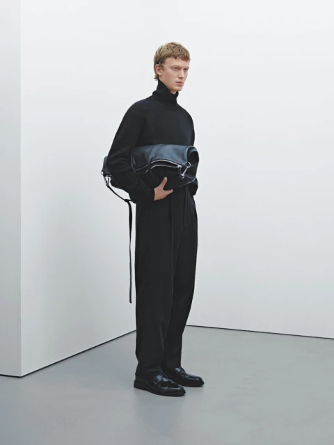 Male model in a black turtleneck and black pants holding a black leather bag in a white photo studio setting.