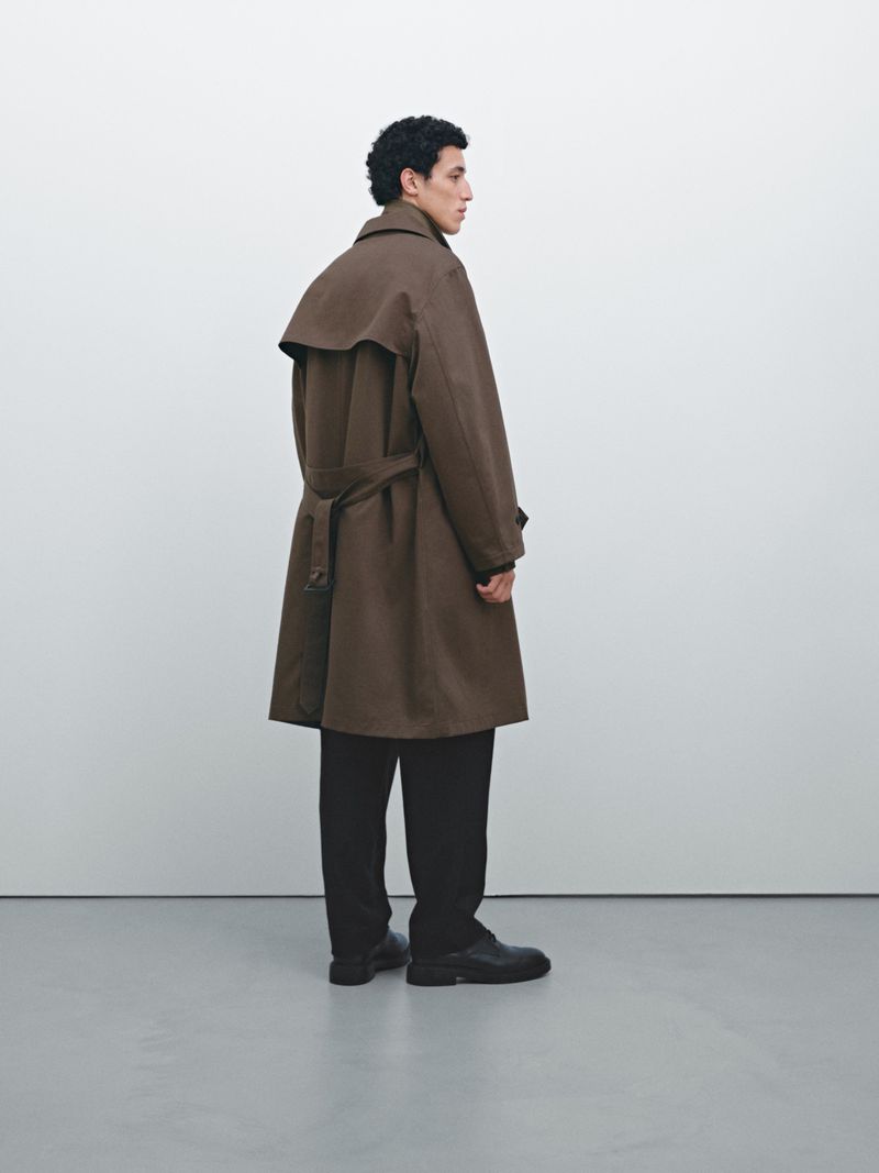 Man in a brown belted trench coat and black pants standing indoors against a white wall in a fashion studio shoot.