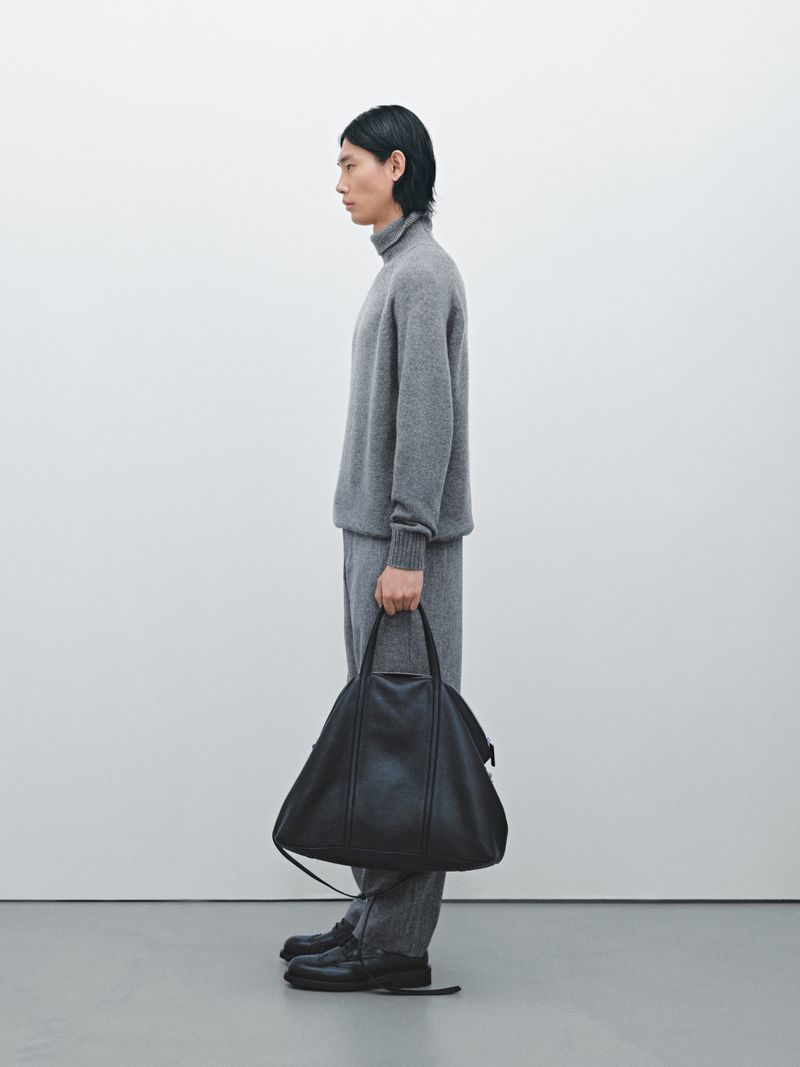 Side profile of an adult male model in a gray turtleneck sweater, gray pants, and black shoes holding a black leather bag.