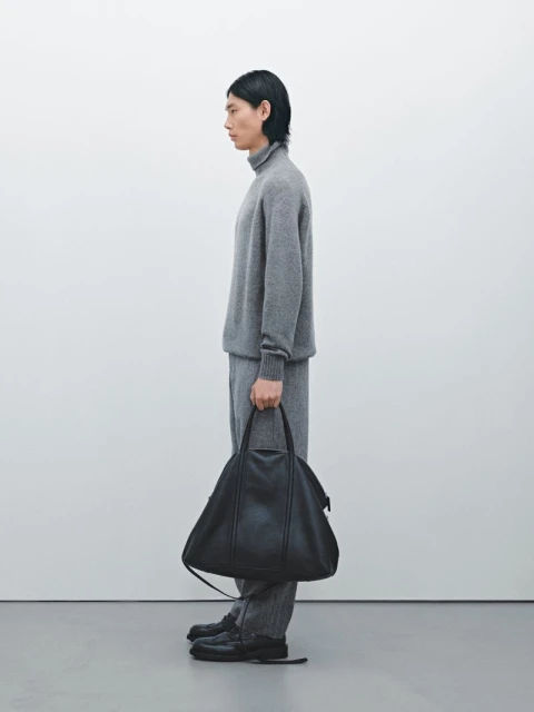 Side profile of an adult male model in a gray turtleneck sweater, gray pants, and black shoes holding a black leather bag.