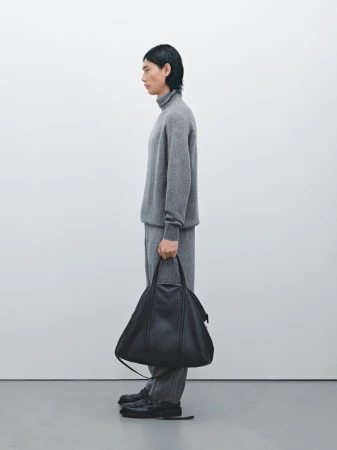 Side profile of an adult male model in a gray turtleneck sweater, gray pants, and black shoes holding a black leather bag.
