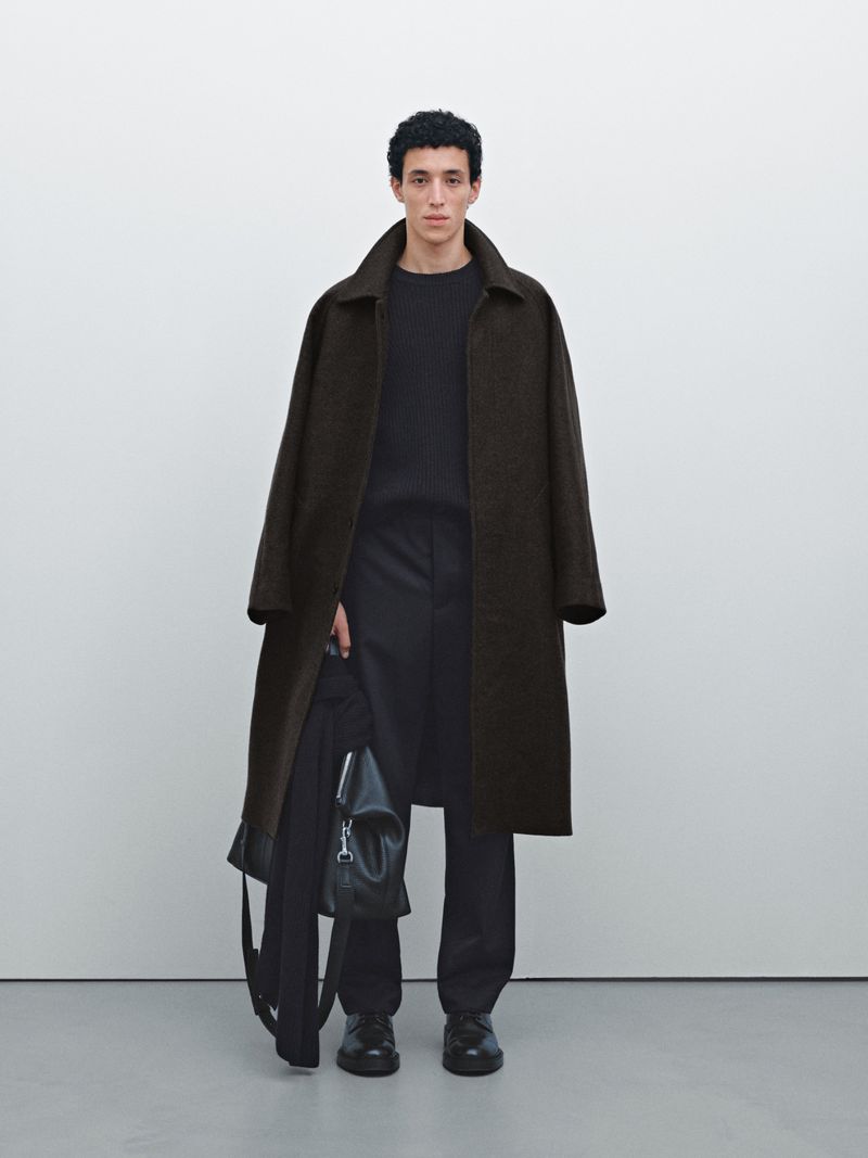 A young man stands in a photo studio wearing a long coat and holding a jacket and bag in his hand, showcasing men's fashion.