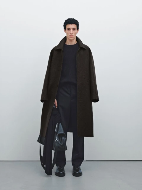 A young man stands in a photo studio wearing a long coat and holding a jacket and bag in his hand, showcasing men's fashion.