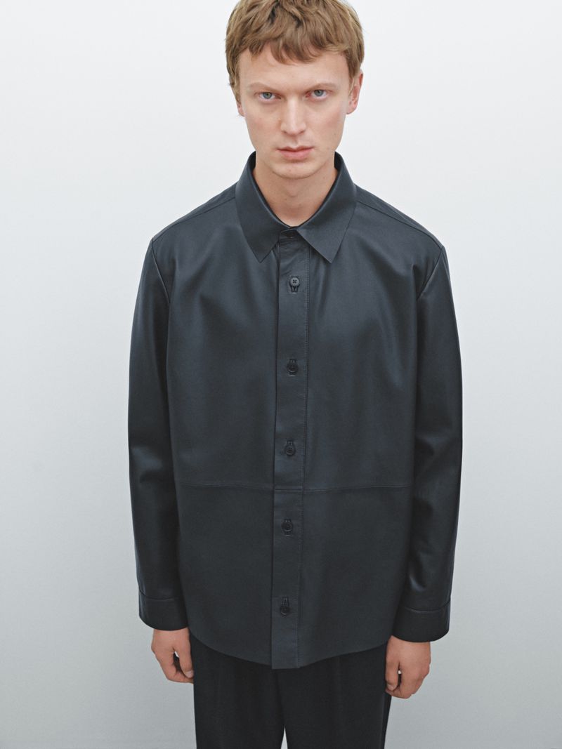 Adult male fashion model in a photo studio wearing a black button-down leather-like shirt and black pants against a white backdrop.
