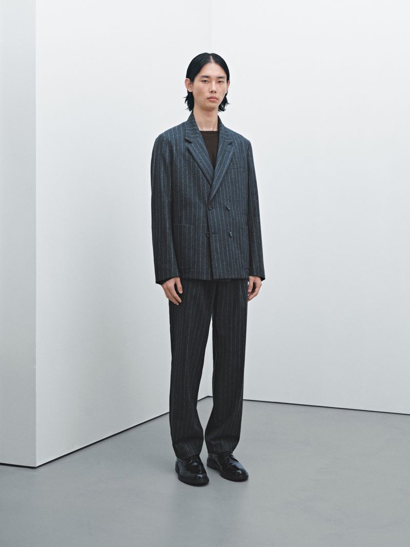 An Asian male model poses in a grey pinstripe suit indoors, standing against a white wall and grey floor with neutral lighting.