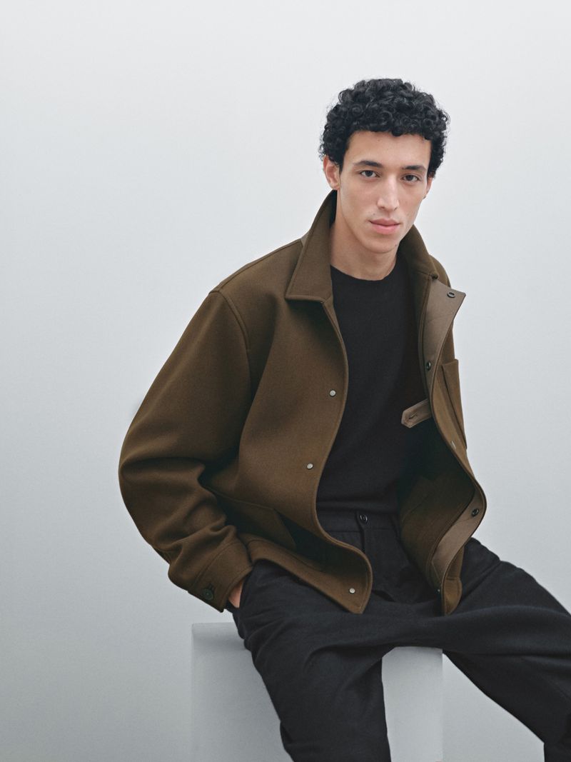 Man wearing a brown collared jacket and black pants sits indoors against a plain white background in a studio setting.