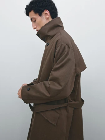 Side profile of a man wearing a high-collar brown belted trench coat against a clean light grey studio background.