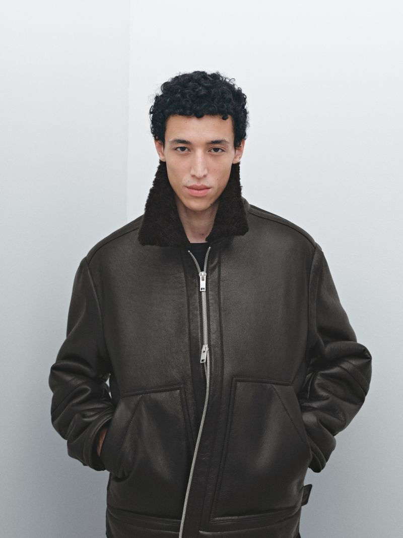 A young adult male poses indoors wearing a brown aviator-style jacket with a dark fur collar against a neutral wall background.