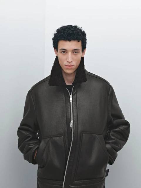 A young adult male poses indoors wearing a brown aviator-style jacket with a dark fur collar against a neutral wall background.