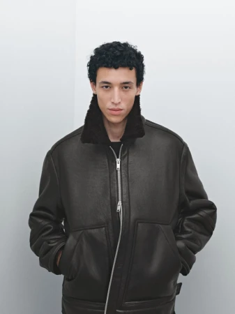 A young adult male poses indoors wearing a brown aviator-style jacket with a dark fur collar against a neutral wall background.