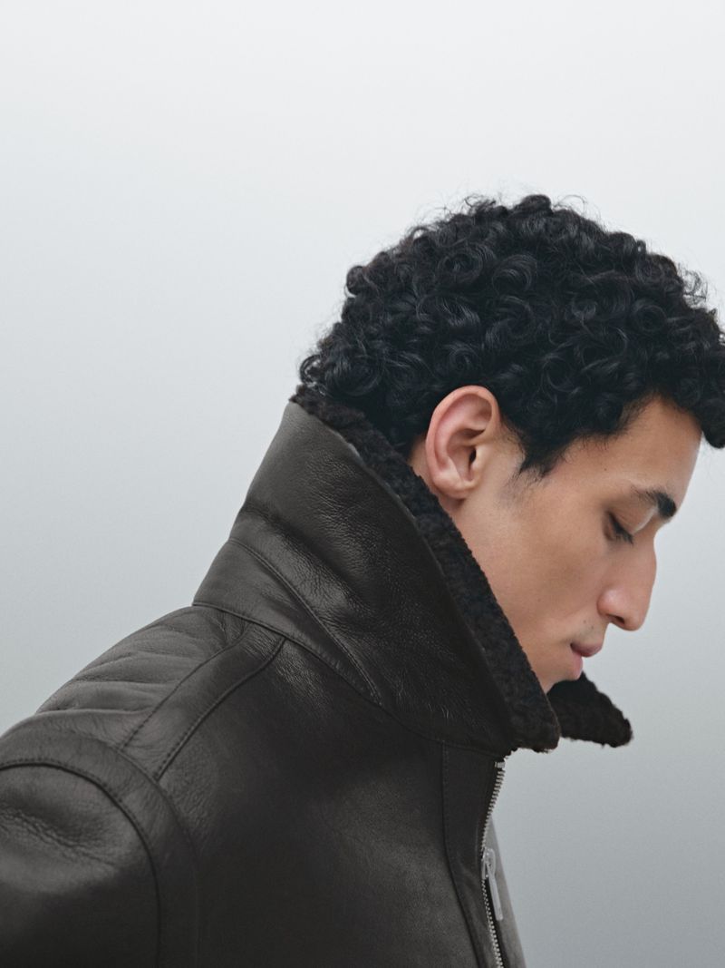 Profile shot of a man with curly hair wearing a brown leather jacket with a shearling collar against a gradient grey background.
