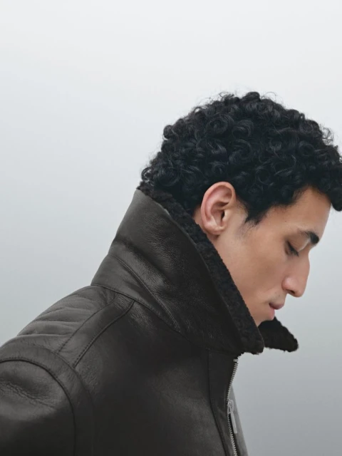 Profile shot of a man with curly hair wearing a brown leather jacket with a shearling collar against a gradient grey background.