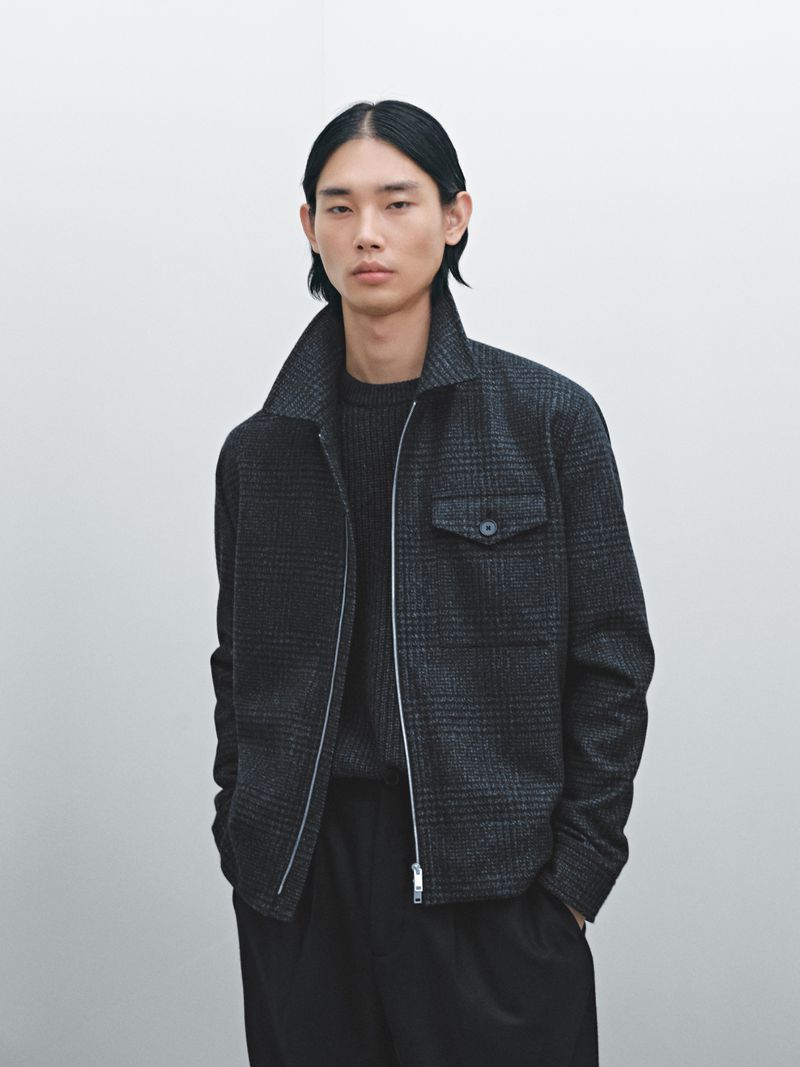 Asian male poses in a photo studio, wearing a grey wool jacket, black trousers, and a knitted wool sweater in a neutral setting.