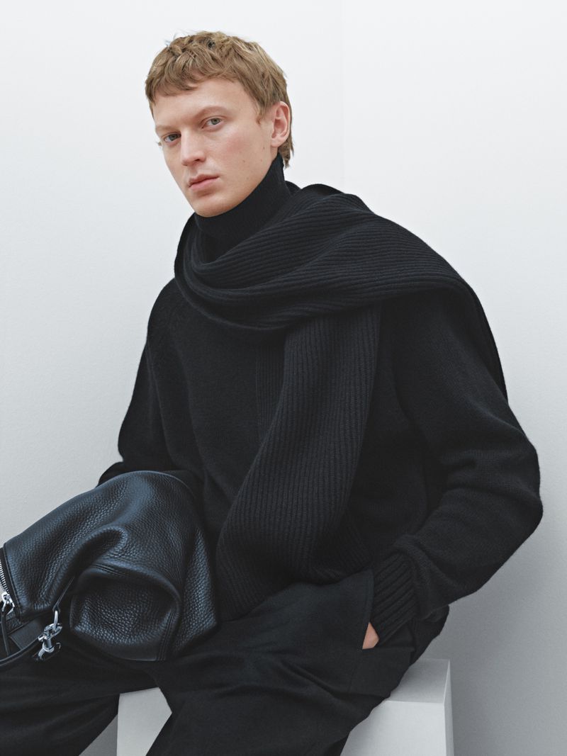 A fair-skinned male fashion model poses seated, wearing black turtleneck sweater and scarf in studio with a bag.