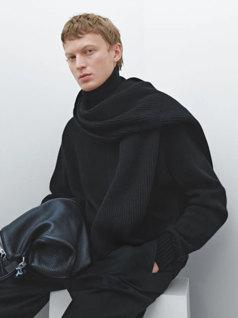 A fair-skinned male fashion model poses seated, wearing black turtleneck sweater and scarf in studio with a bag.