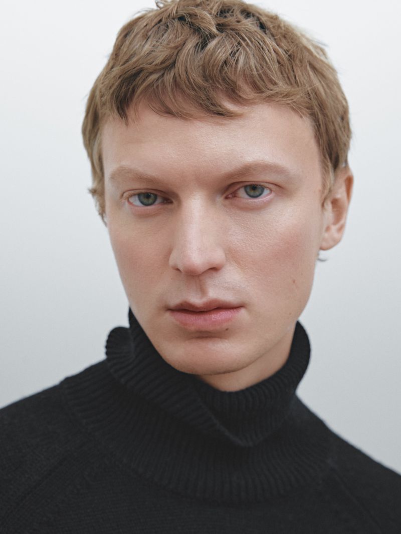Headshot of a fair-skinned male wearing a black turtleneck sweater against a white background, creating a simple yet striking portrait.