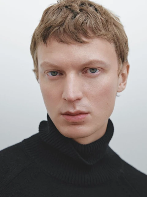 Headshot of a fair-skinned male wearing a black turtleneck sweater against a white background, creating a simple yet striking portrait.