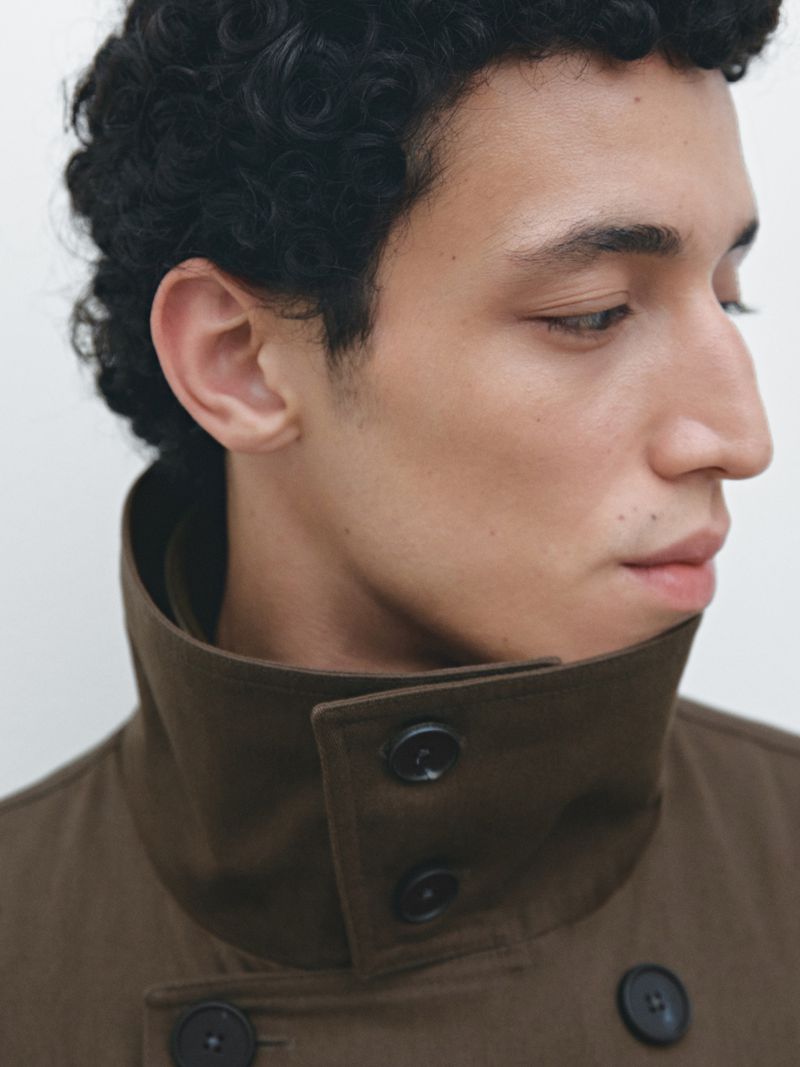 Close-up of a young adult male with curly dark hair, wearing a brown collared jacket, against a neutral white background.