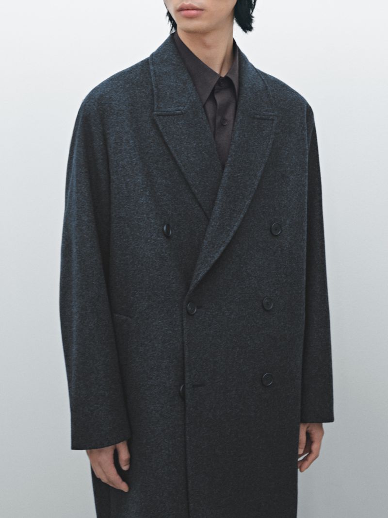 An adult male stands indoors wearing a grey wool double-breasted overcoat with a dark brown collared shirt against a white backdrop.