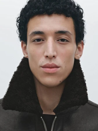 Portrait of a young adult male with dark curly hair, wearing a brown leather jacket with a shearling collar against a plain white backdrop.