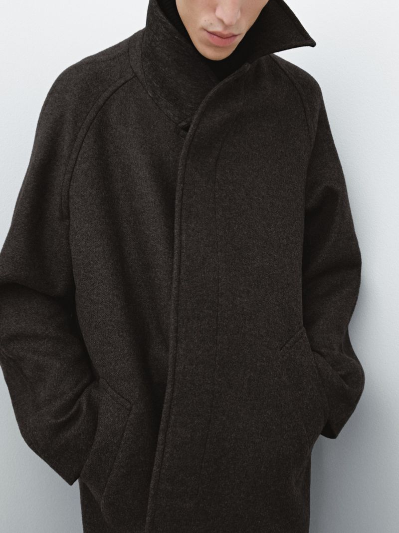 Close-up of a male fashion model showcasing a charcoal grey wool overcoat, hands in pockets, against a plain grey studio backdrop.