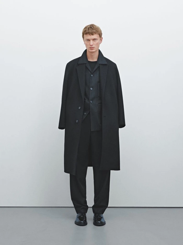 Model Jonas Glöer standing in an all-black tailored overcoat and matching trousers against a clean white studio backdrop.