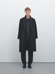 Model Jonas Glöer standing in an all-black tailored overcoat and matching trousers against a clean white studio backdrop.