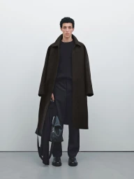 Model Walid Fiher wearing a long dark brown overcoat, black crewneck knit, and black trousers against a neutral background.