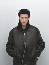 Model wearing a dark leather bomber jacket with a shearling collar against a plain white studio background.