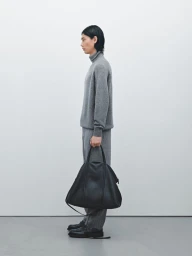 Profile view of a male model wearing a grey turtleneck sweater and matching trousers, holding a large black leather bag.