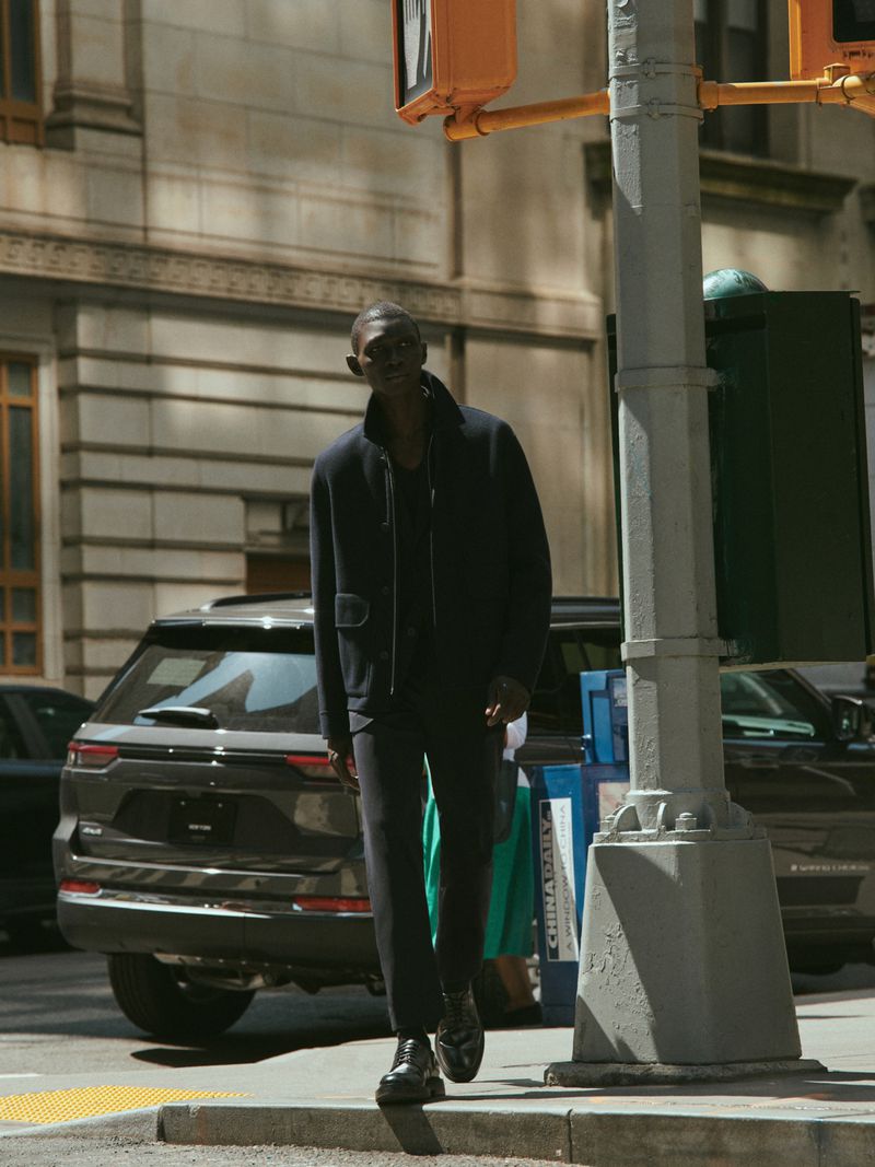 An African American man in a navy jacket and pants crosses a city street at a crosswalk, with cars and buildings in the background.