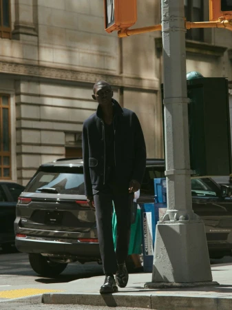 An African American man in a navy jacket and pants crosses a city street at a crosswalk, with cars and buildings in the background.