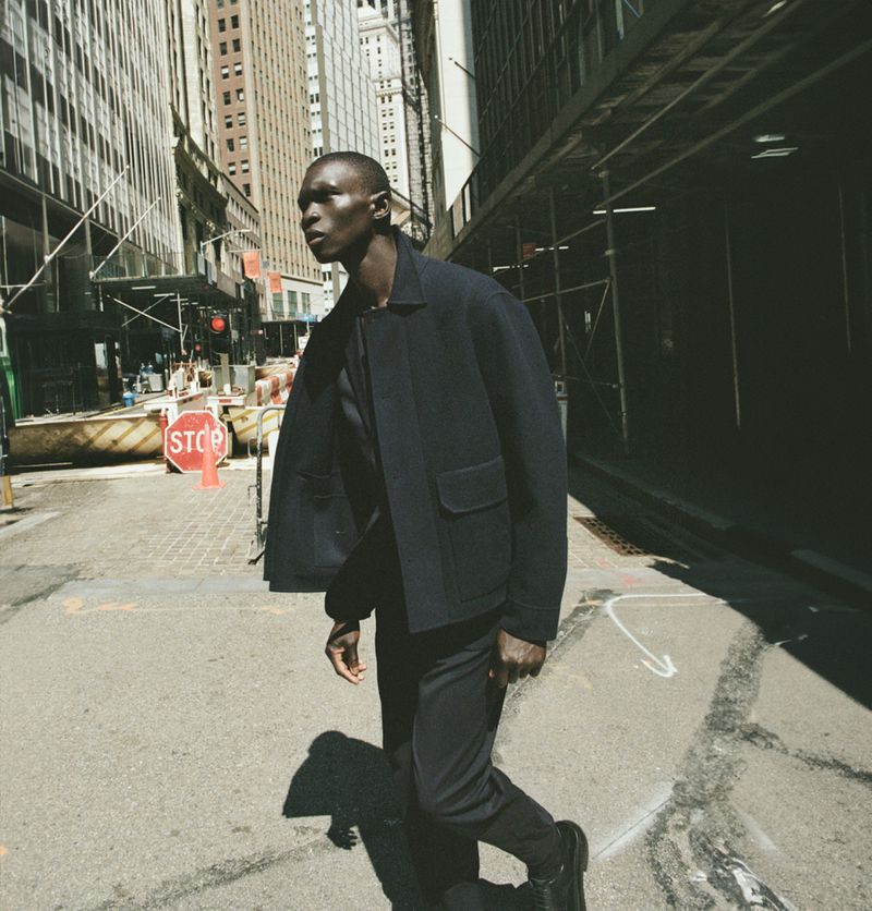 An adult man wearing a dark jacket and pants walks down a city street with tall buildings in the background and a stop sign nearby.