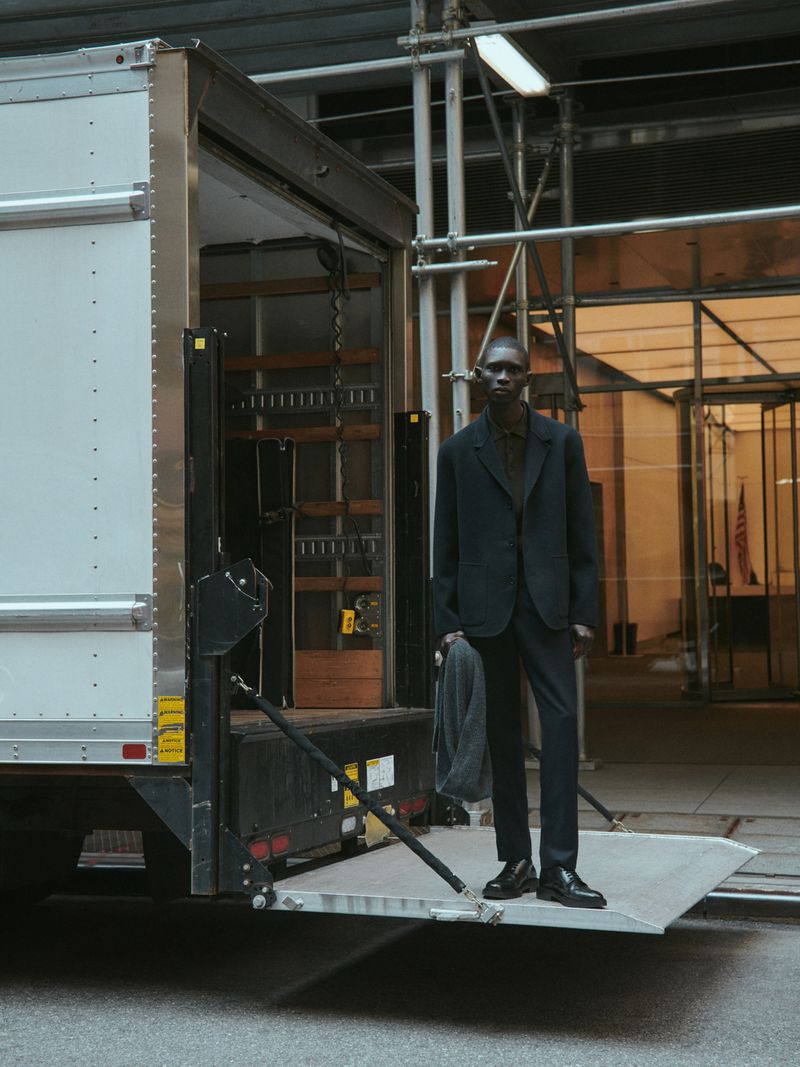 An adult African-American male stands on a truck lift wearing a dark suit in an urban setting with scaffolding visible.