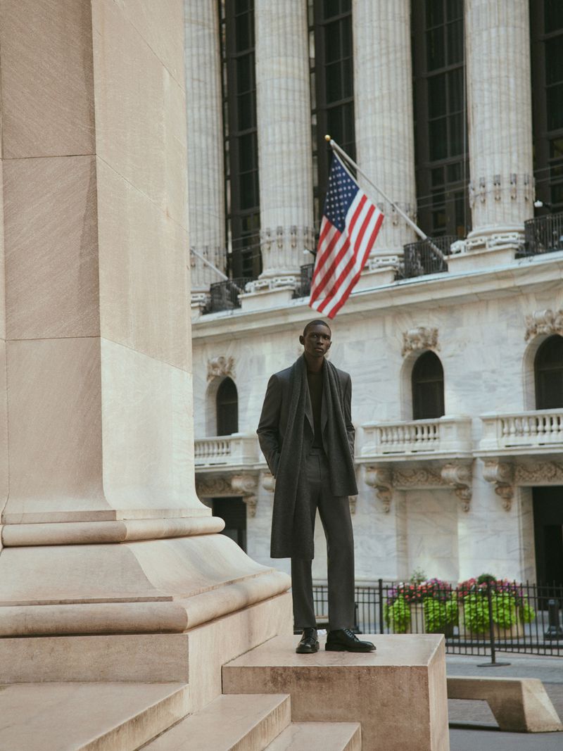 A Black man stands on stone steps in a grey suit and overcoat outside a building with an American flag, creating a professional fashion editorial.