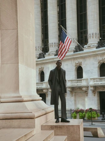 A Black man stands on stone steps in a grey suit and overcoat outside a building with an American flag, creating a professional fashion editorial.