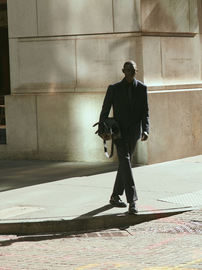 An African-American adult male in a suit is walking on a city sidewalk carrying a bag. The building in the background is made of stone.