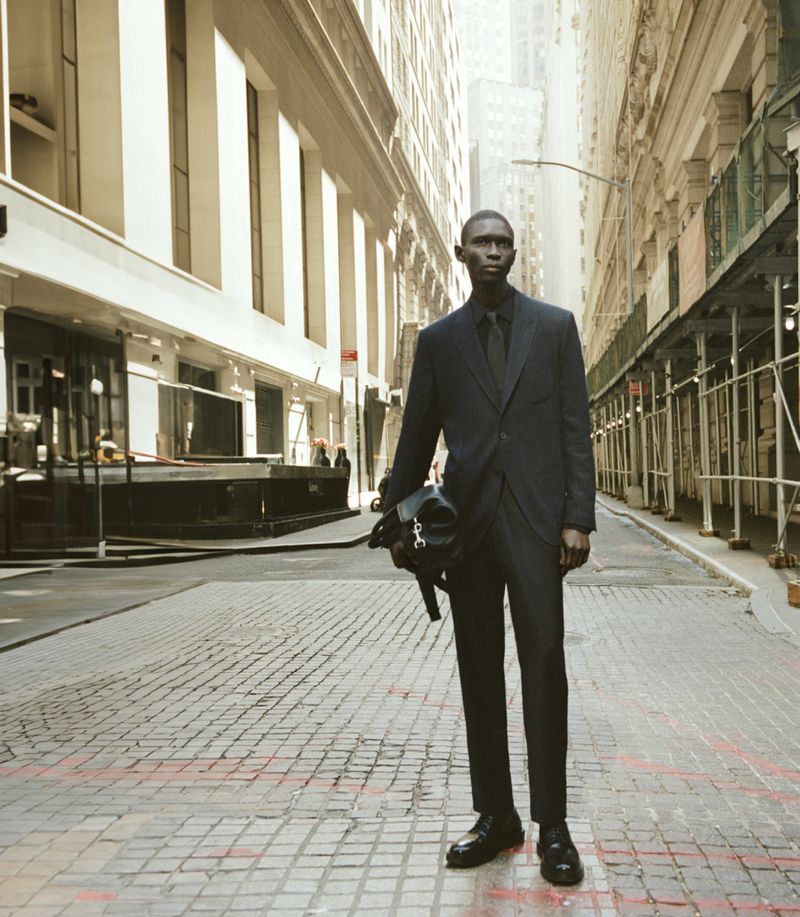 Fashion shot of an adult Black male in a business suit, holding a soft bag, posing in an urban environment with tall buildings.