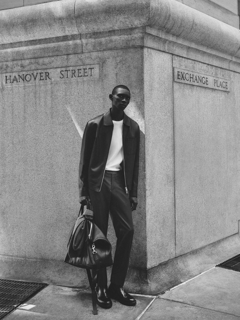 Black and white shot of a Black man leaning against a building corner on Hanover Street, carrying a backpack in an urban setting.