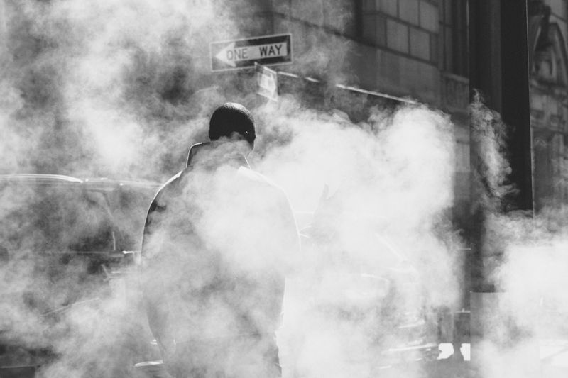 Monochrome shot of a person walking on a city street filled with steam, with a One Way sign overhead and architecture in the background.