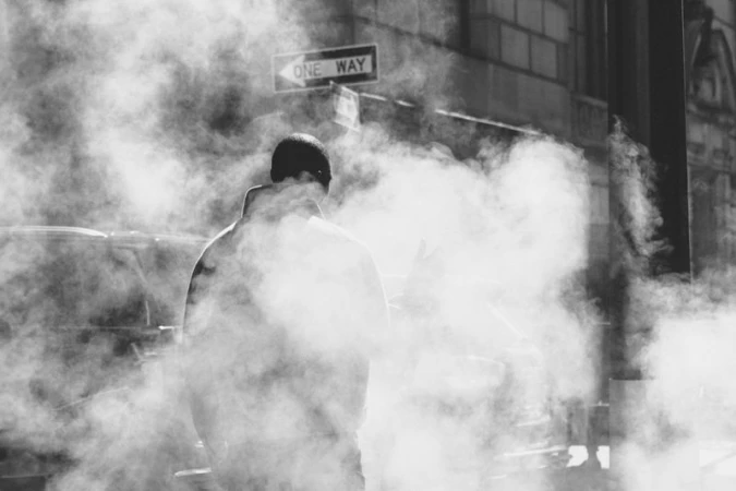 Monochrome shot of a person walking on a city street filled with steam, with a One Way sign overhead and architecture in the background.