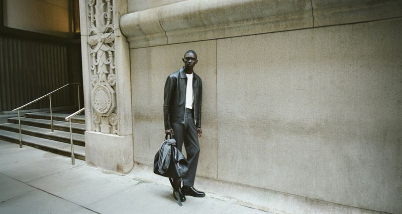 An adult male in an outdoor urban setting wearing a leather jacket, white t-shirt, grey slacks, and black boots with a black carry bag.