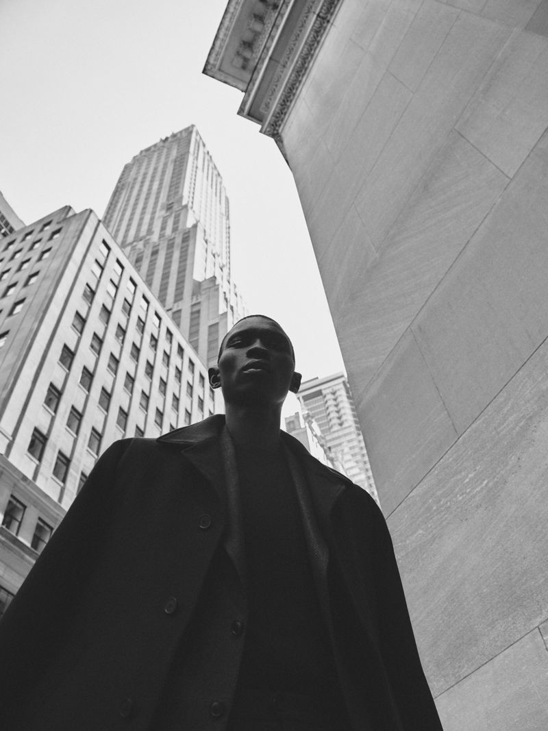 Black and white portrait of a man in a coat standing in a city street, looking up at high-rise buildings and a partly cloudy sky.