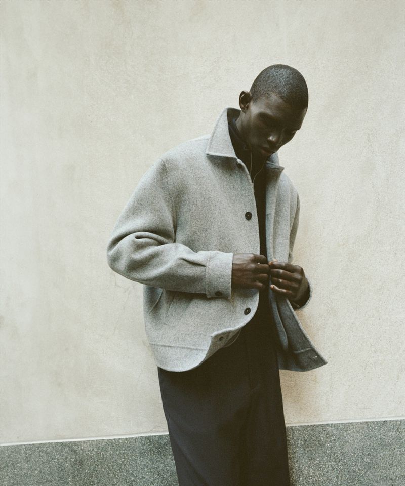 An African American man models a grey jacket and dark pants in a studio setting. The man buttons his jacket in a fashion shoot.