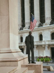 Model Fernando Cabral in a charcoal tailored coat and trousers posing on stone steps before classical columns and an American flag.