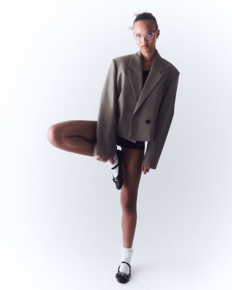 Woman in oversized blazer and glasses poses in a studio. She is standing on one leg, wearing black ballet flats with white socks.