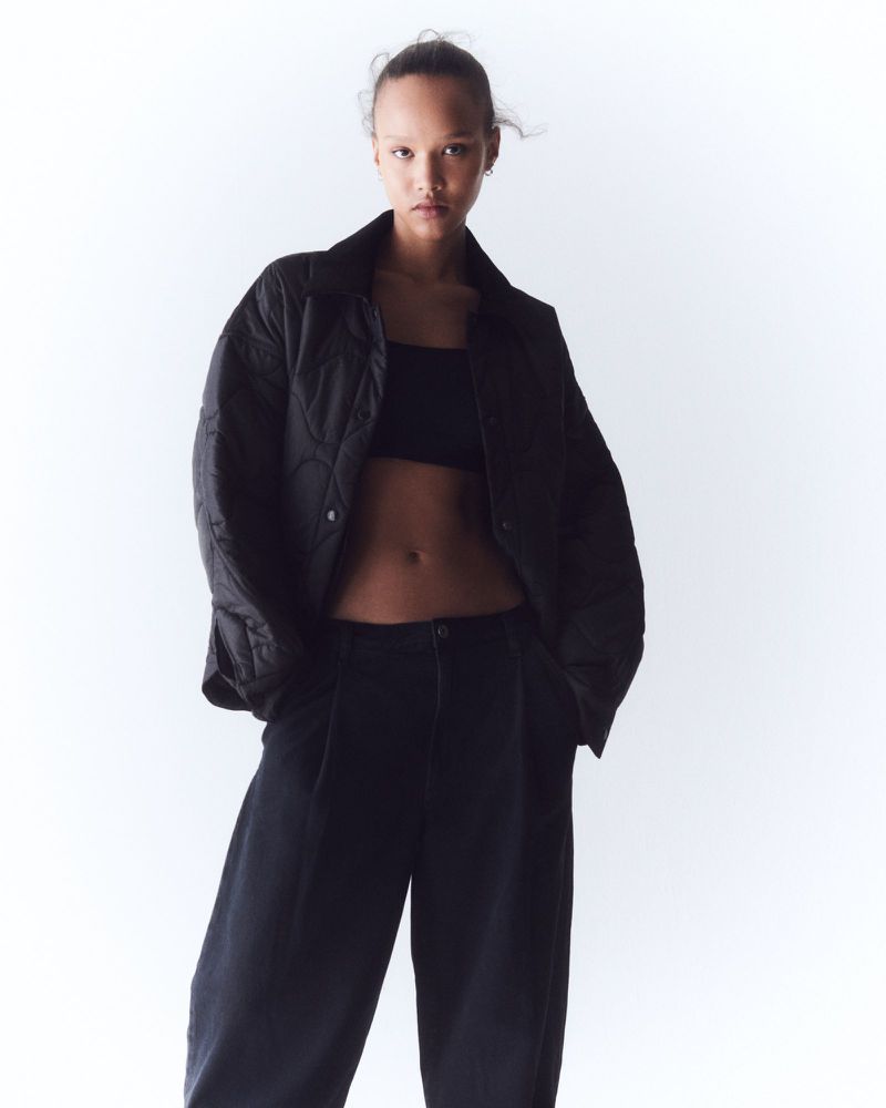 Fashion portrait of a woman in a black jacket and pants against a white background, looking directly at the camera with a neutral expression.